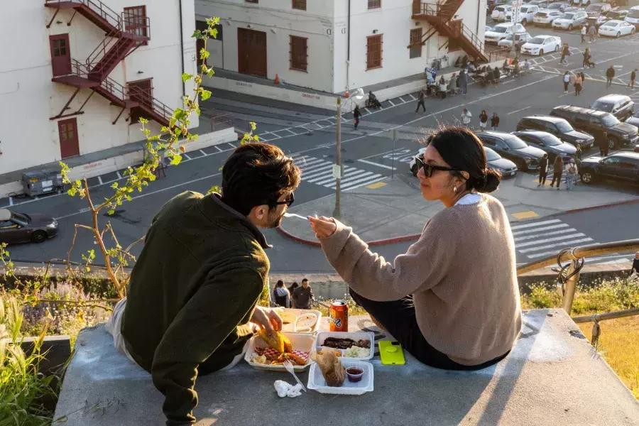 Um casal janta ao ar livre no Fort Mason Center, em São Francisco. A mulher alimenta seu companheiro com um gostinho de comida.