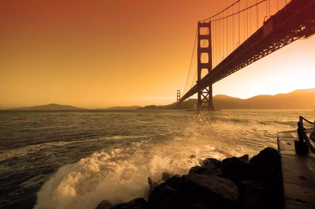 Waves crash near Fort Point beneath the Golden Gate Bridge at sunset.