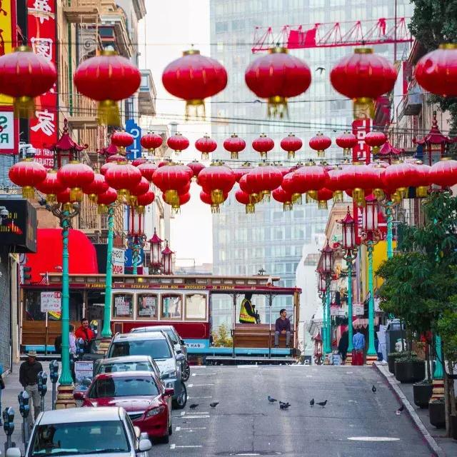 Uma rua montanhosa na Chinatown de São Francisco é retratada com lanternas vermelhas penduradas e um bonde passando.